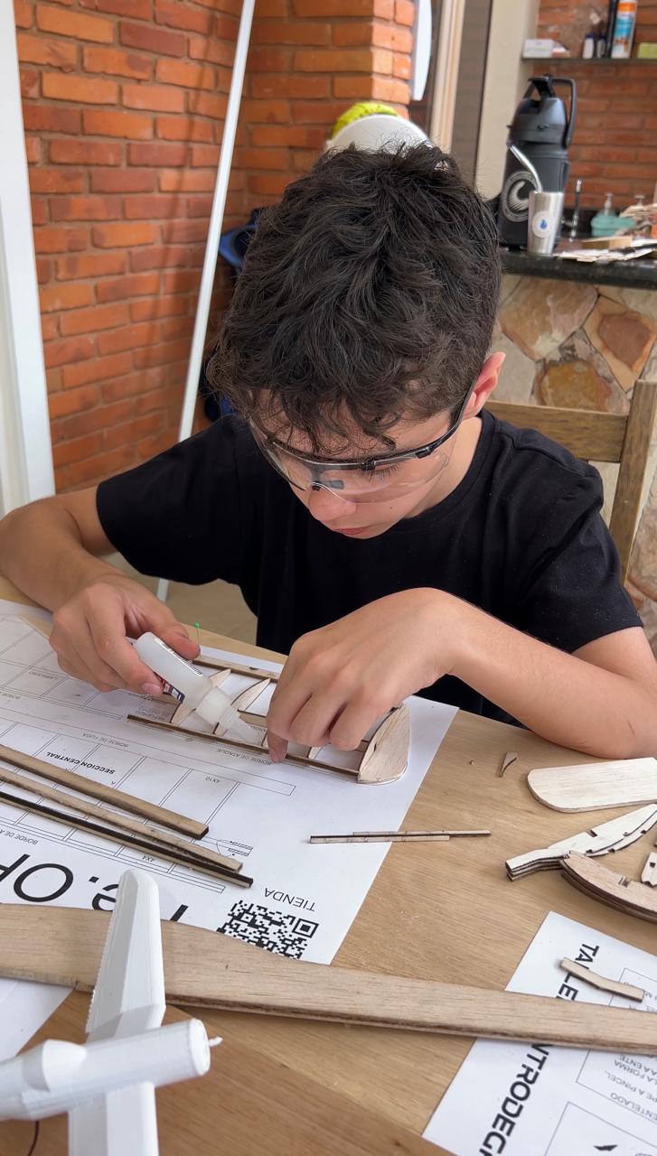 Chico con lentes de seguridad pegando las piezas de balsa de un ala sobre el plano del aeromodelo